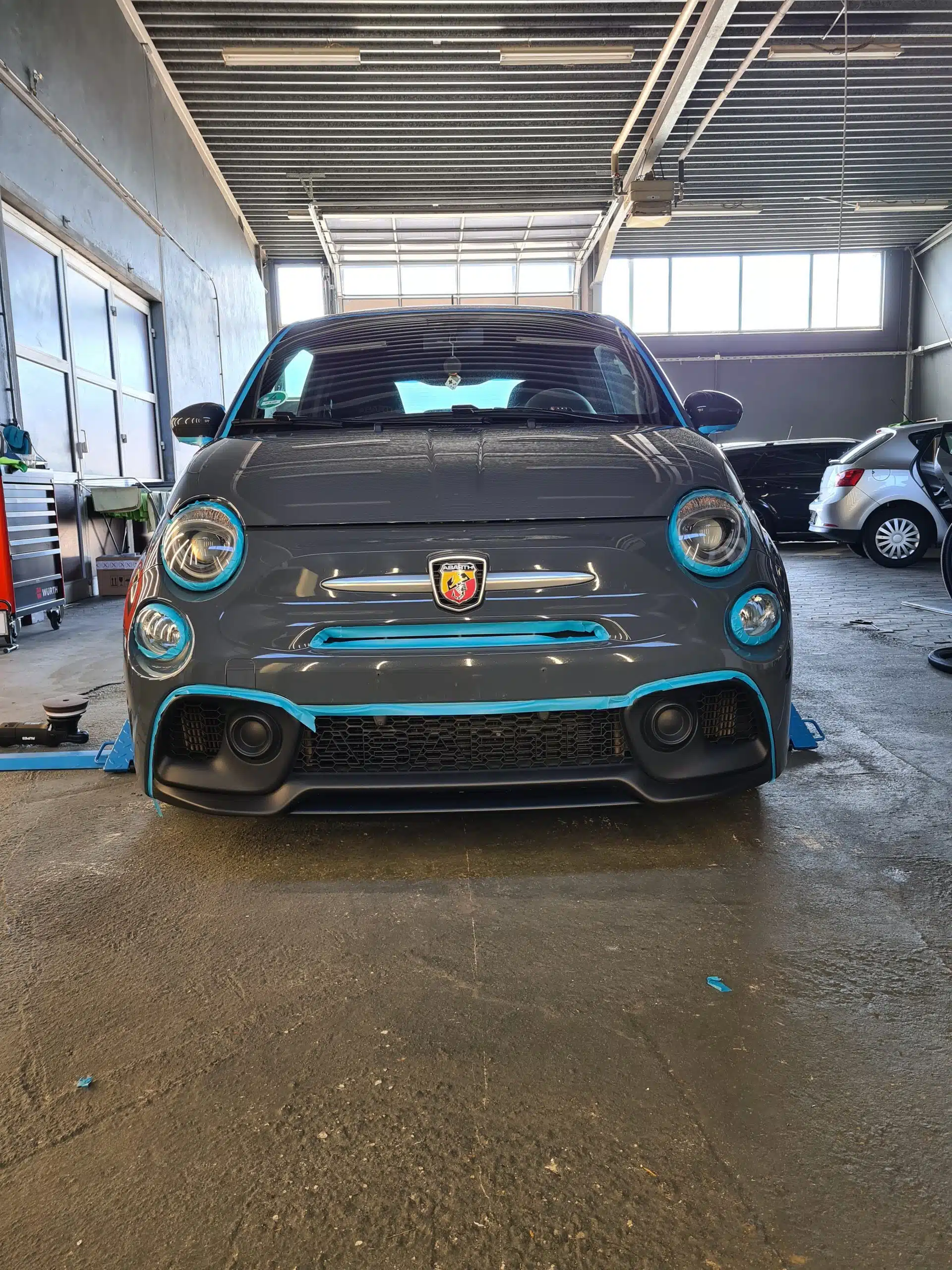 Grauer Abarth 595, für die Aufbereitung vorbereitet und mit blauem Abdeckband geschützt, in einer Werkstatthalle.