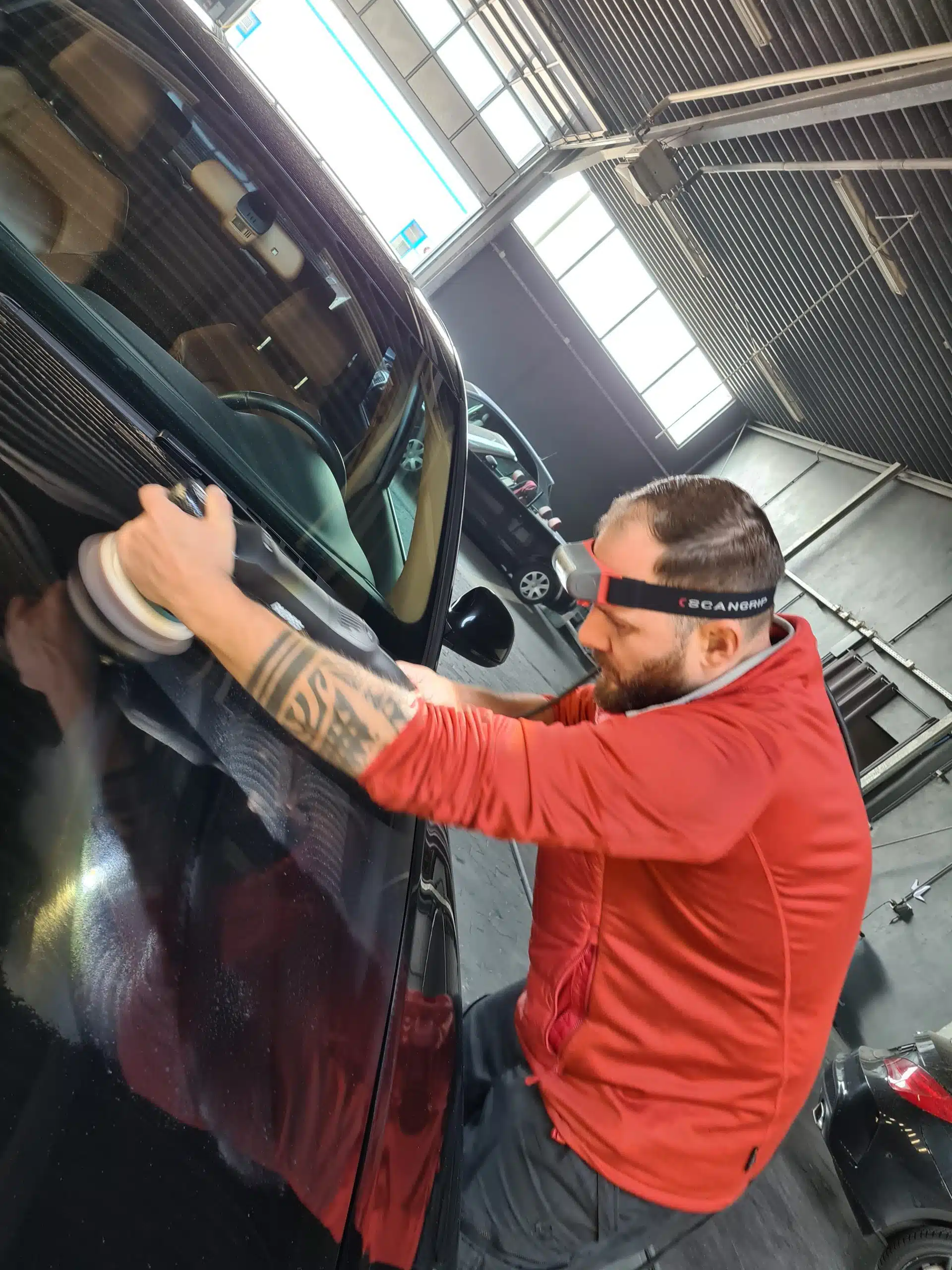 Fahrzeugaufbereiter poliert den Lack eines schwarzen Autos mit einer Poliermaschine in einer Werkstatthalle.