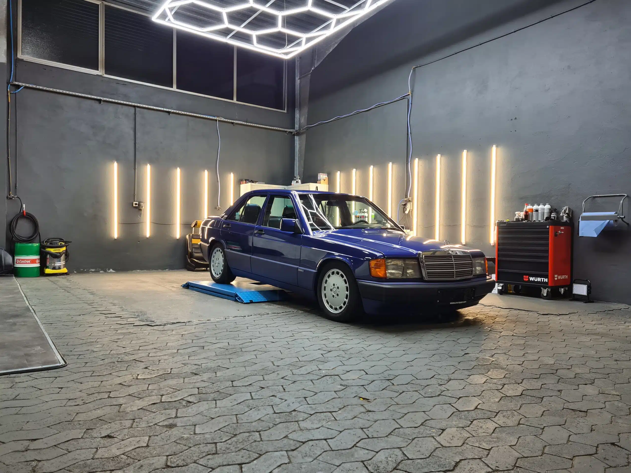 Blauer Mercedes 190E auf einer niedrigen Aufbereitungsplattform, in Halle mit vertikalen LED-Lichtleisten.