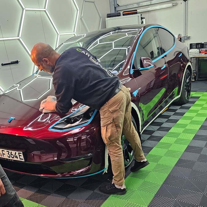 Techniker bereitet einen burgundroten Tesla für die Lackaufbereitung vor, mit blauem Abdeckband geschützt, im Aufbereitungsraum des Car & Clean Centers.