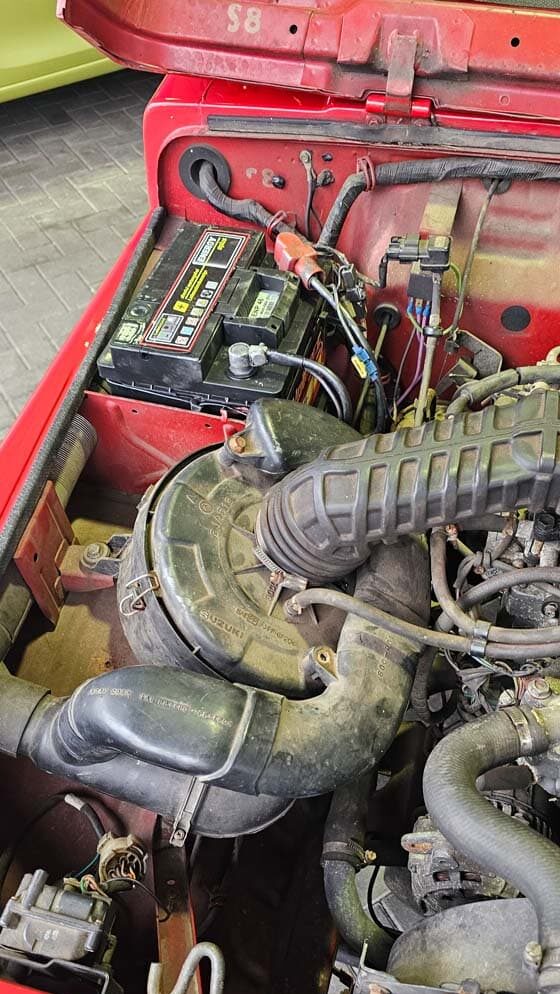 Verschmutzter Motorraum eines roten Fahrzeugs vor der Reinigung, mit sichtbarer Batterie, Luftfiltergehäuse und mehreren Schläuchen.