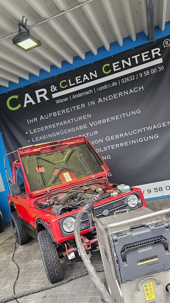 Roter Suzuki-Geländewagen mit geöffnetem Motorraum vor dem Car & Clean Center in Andernach während einer Motorraum-Aufbereitung.