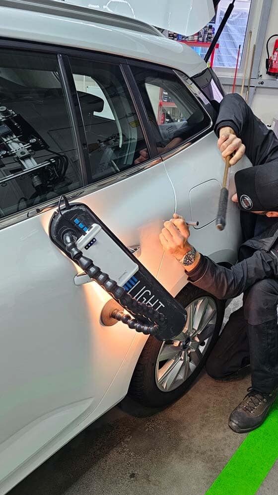 Fachgerechte Dellenentfernung an einem weißen Fahrzeug mit Spezialwerkzeug und Ausbeullampe.