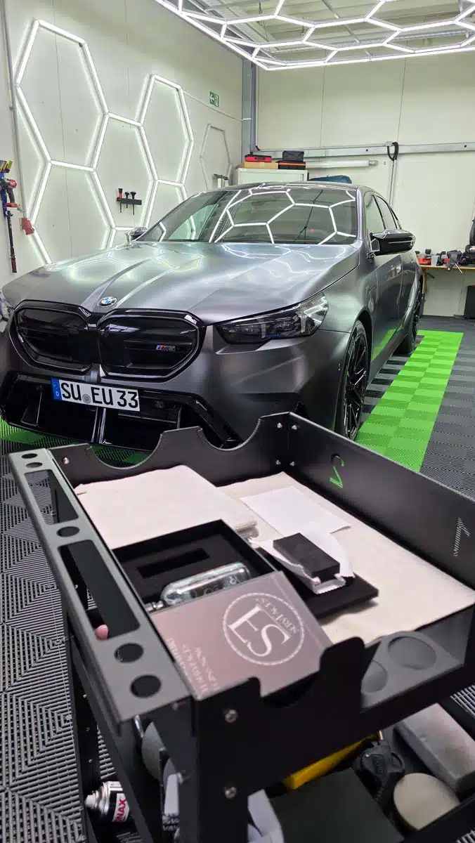 Mattschwarzer BMW in moderner Detailing-Halle, im Vordergrund ein Vorbereitungstisch mit Beschichtungs-Equipment.